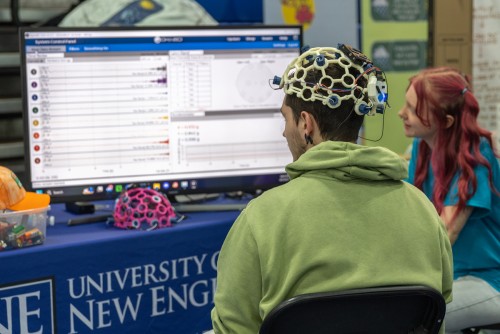 A guest at a health fair wears a brain wave helmet while a neuroscience student monitors the data showing up on a large monitor