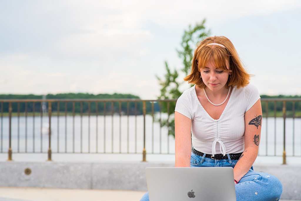 A student sites in front of the water on a bench working on a laptop