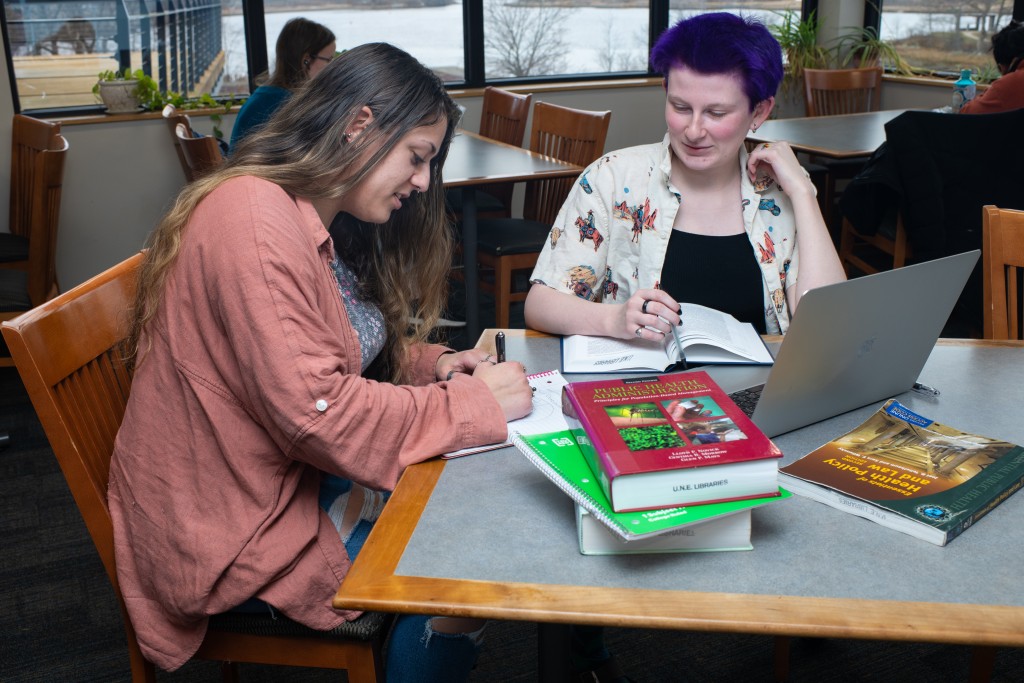 Two U N E study together with a stack of textbooks and a laptop