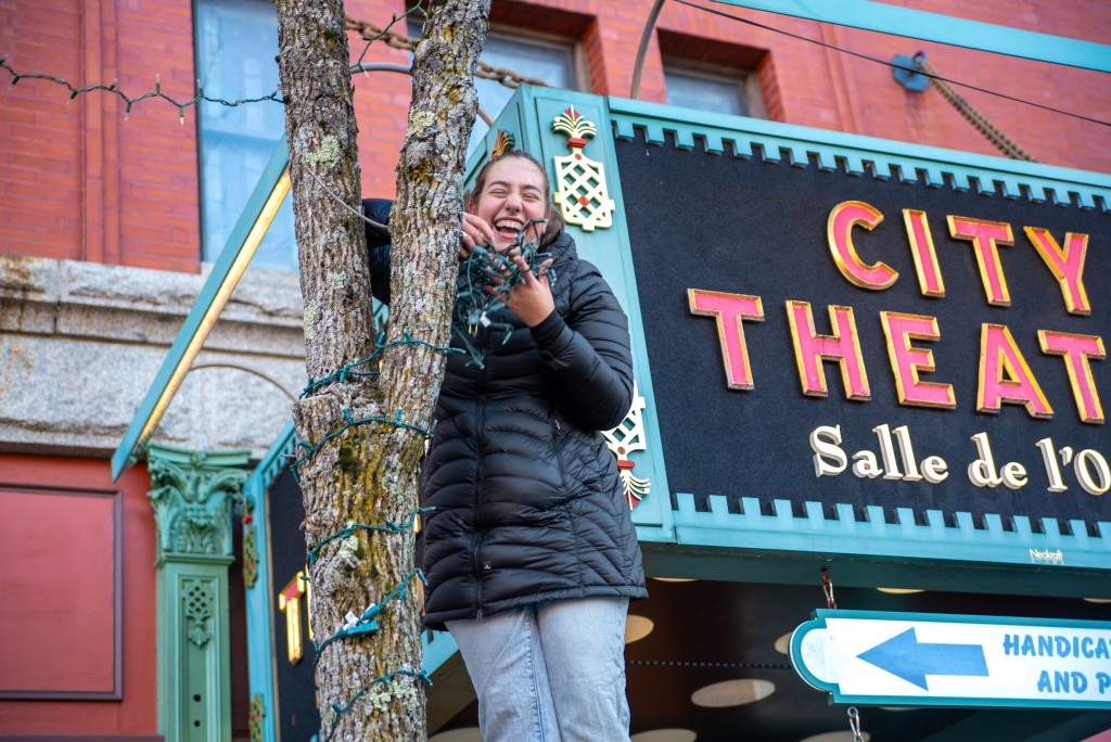 A female student wraps lights around the trunk of a tree in downtown Biddeford