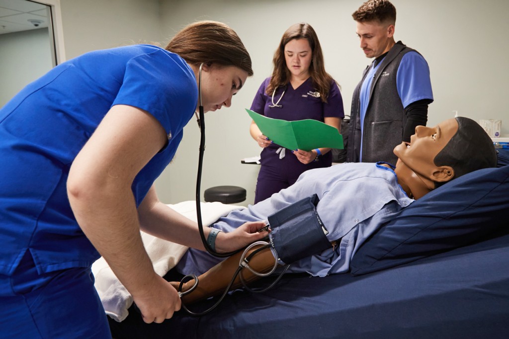 A P A student practices checking blood pressure on a patient simulator while two other students discuss the patient's health history