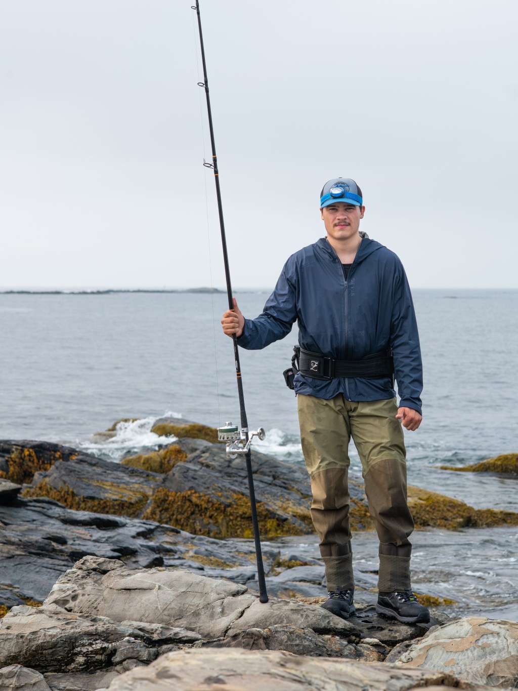 A UNE student poses in front of Saco Bay holding a fishing rod