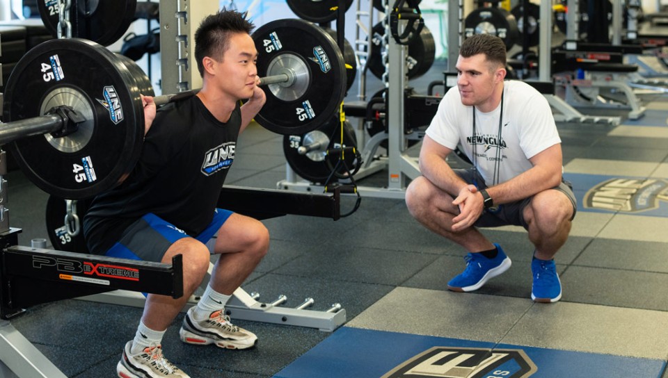 A student coaches another student performing squats with a barbell on their back