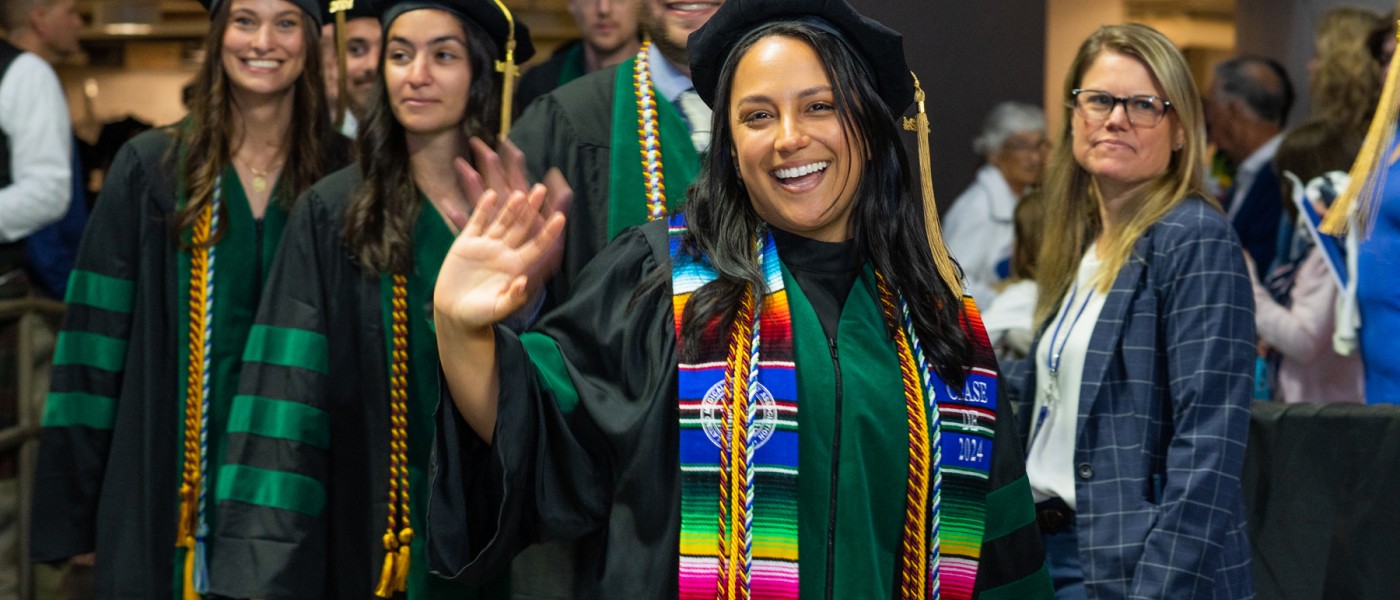 A UNE graduate wearing doctoral regalia waves walking into the ceremony